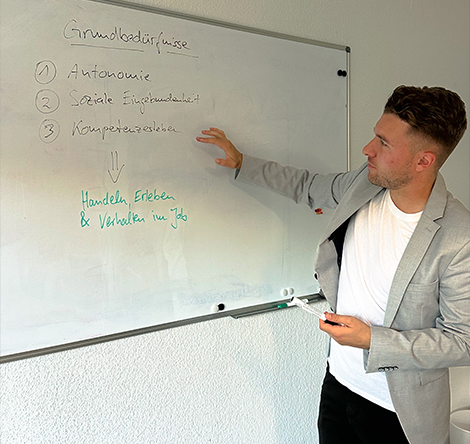 Philipp Romahn steht vor einer Tafel auf der Grundbedürfnisse aufgeschrieben sind. Er zeigt mit dem Finger auf den Text Kompetenzen, der auf der Tafel steht.