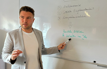 Philipp Romahn steht links im Bild und rechts von ihm ist die Tafel. Er schaut in den Raum und zeigt mit dem Stift auf die Tafel.